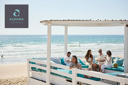 Mehrere Personen sitzen in einer Strandbar, im Hintergrund Strand und Meer